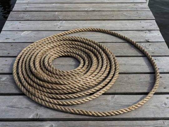 Paksu hamppuköysi kiertyneenä harmaantuneelle puulaiturille niin että köyden kaari osoittaa oikeaan reunaan